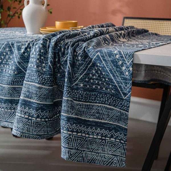 Cotton-linen tablecloth with navy geometric print and tassel trim, styled on a wooden table with white vase and warm-toned wall.