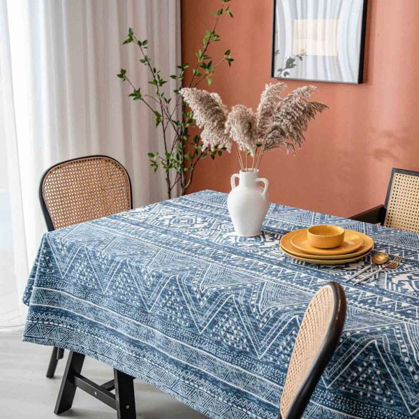 Cotton-linen tablecloth with navy geometric print and tassel trim, styled on a wooden table with white vase and warm-toned wall.
