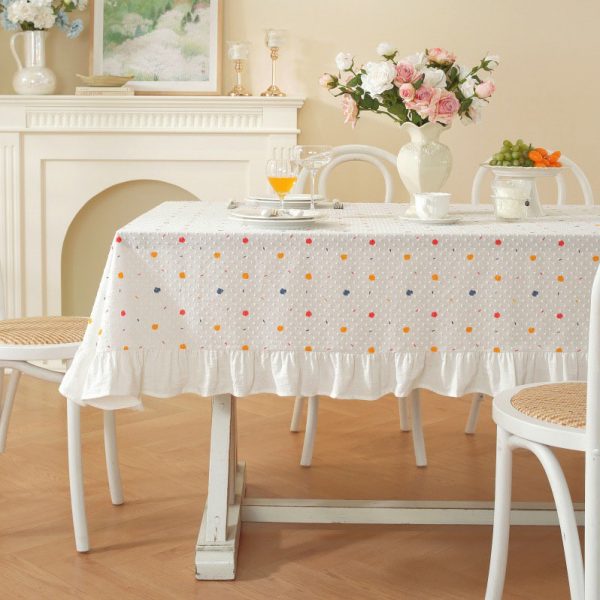 White cotton tablecloth with colorful polka dots and ruffled trim, styled on a dining table with light wooden chairs and soft natural lighting.