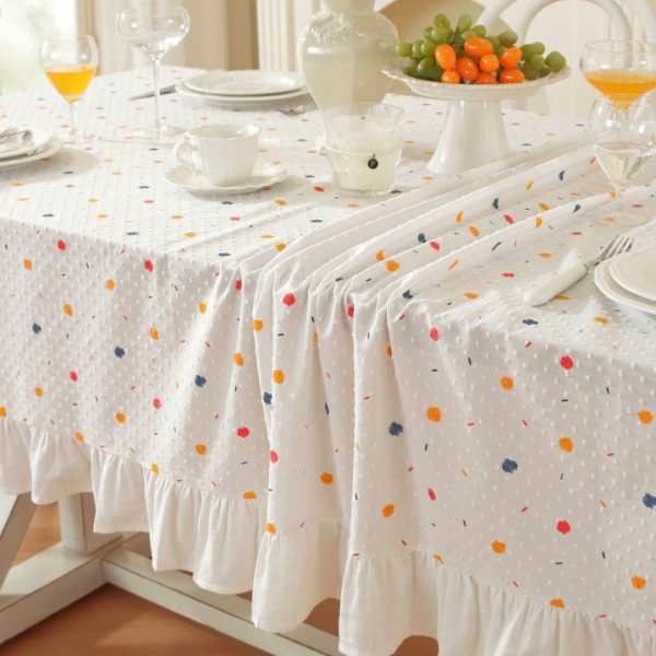 White cotton tablecloth with colorful polka dots and ruffled trim, styled on a dining table with light wooden chairs and soft natural lighting.