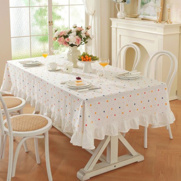 White cotton tablecloth with colorful polka dots and ruffled trim, styled on a dining table with light wooden chairs and soft natural lighting.