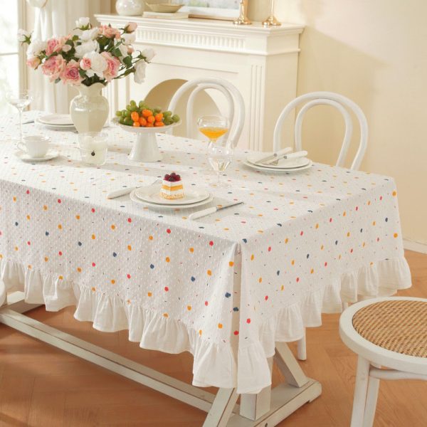 White cotton tablecloth with colorful polka dots and ruffled trim, styled on a dining table with light wooden chairs and soft natural lighting.