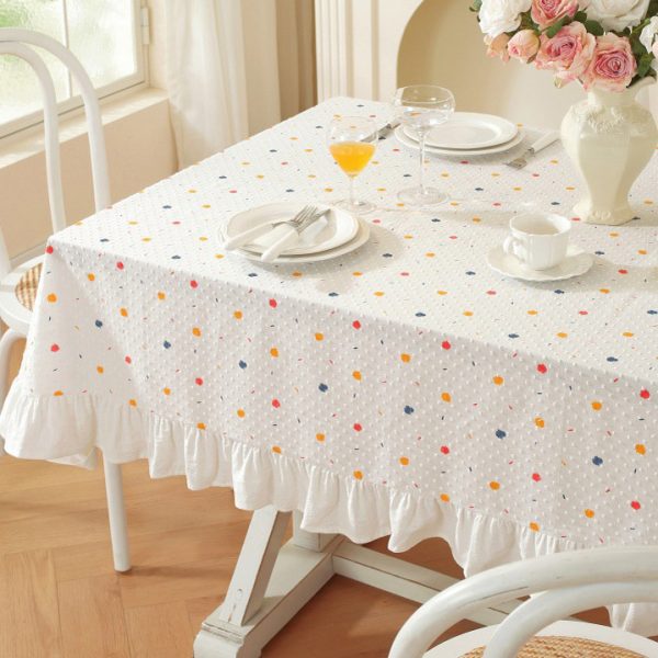 White cotton tablecloth with colorful polka dots and ruffled trim, styled on a dining table with light wooden chairs and soft natural lighting.