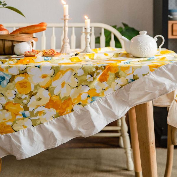 Yellow floral cotton tablecloth with white ruffled trim, styled on a rustic table for spring or cottagecore dining. Features soft fabric, bright flower print, and elegant edge detail.