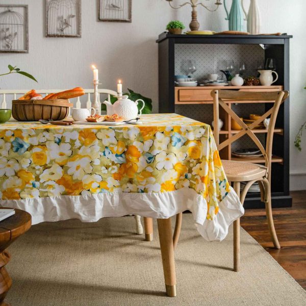 Yellow floral cotton tablecloth with white ruffled trim, styled on a rustic table for spring or cottagecore dining. Features soft fabric, bright flower print, and elegant edge detail.