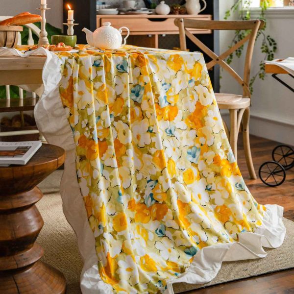Yellow floral cotton tablecloth with white ruffled trim, styled on a rustic table for spring or cottagecore dining. Features soft fabric, bright flower print, and elegant edge detail.