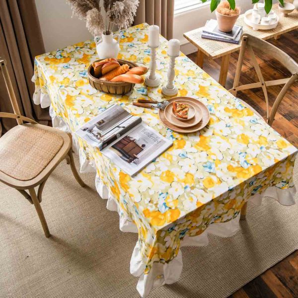 Yellow floral cotton tablecloth with white ruffled trim, styled on a rustic table for spring or cottagecore dining. Features soft fabric, bright flower print, and elegant edge detail.