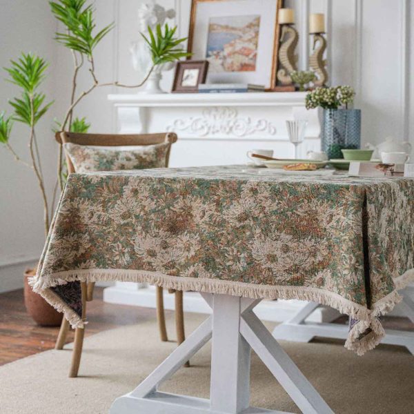 A green jacquard tablecloth with oil painting-style floral design and white tassel edges, styled on a white table with wooden chairs for a warm rustic look.