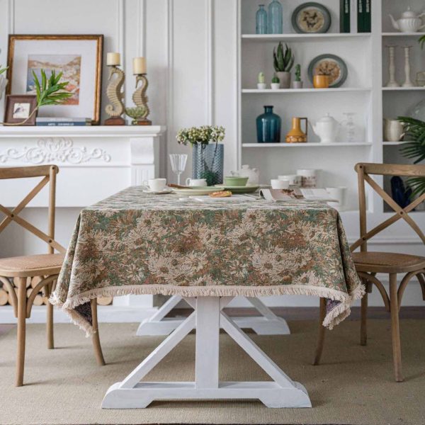 A green jacquard tablecloth with oil painting-style floral design and white tassel edges, styled on a white table with wooden chairs for a warm rustic look.
