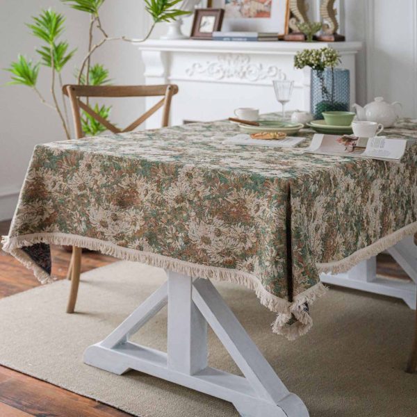 A green jacquard tablecloth with oil painting-style floral design and white tassel edges, styled on a white table with wooden chairs for a warm rustic look.