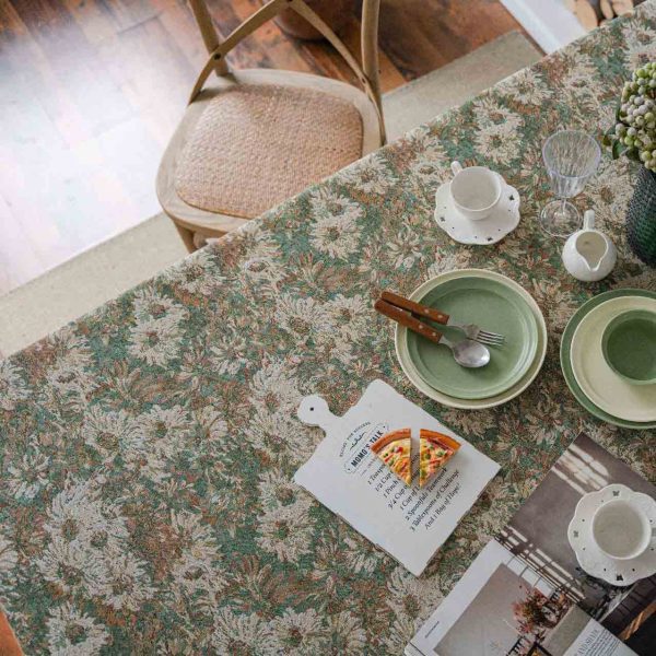 A green jacquard tablecloth with oil painting-style floral design and white tassel edges, styled on a white table with wooden chairs for a warm rustic look.