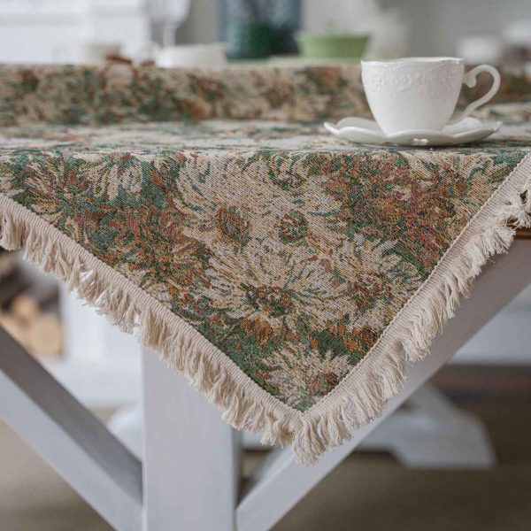 A green jacquard tablecloth with oil painting-style floral design and white tassel edges, styled on a white table with wooden chairs for a warm rustic look.