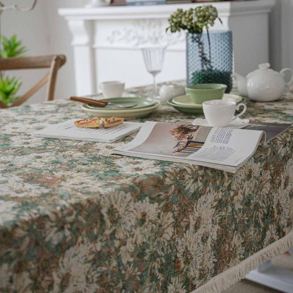 A green jacquard tablecloth with oil painting-style floral design and white tassel edges, styled on a white table with wooden chairs for a warm rustic look.
