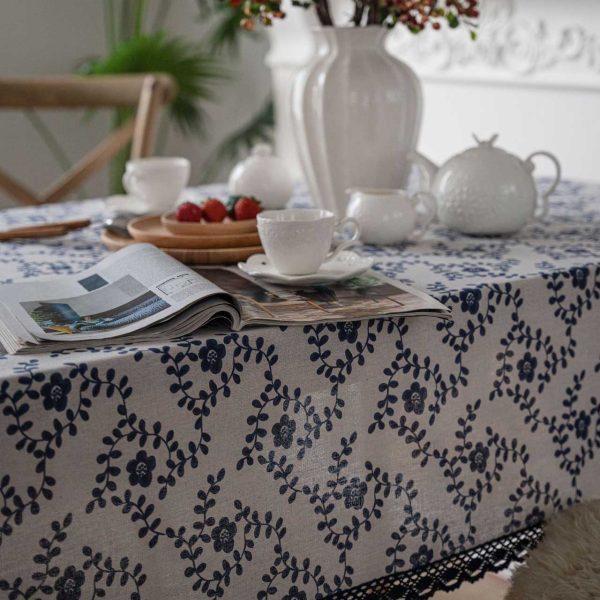 A hand-painted blue vine floral cotton linen tablecloth with navy lace trim, styled on a rustic wooden table with minimalist dinnerware and greenery.